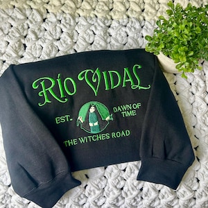 May include: Black long-sleeve shirt with green embroidered text that reads "Rio Vidal Est. The Witches Road Dawn of Time". The design features a green silhouette of a woman in a hooded cloak.