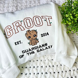 May include: White sweatshirt with brown embroidered text "GROOT" and a cartoon image of Groot from Guardians of the Galaxy. The text "EST. 2014" and "GUARDIANS OF THE GALAXY" are also embroidered in black.