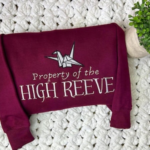 May include: A maroon sweatshirt with the text "Property of the HIGH REEVE" in white script. Above the text is a white origami crane. The sweatshirt is folded on a white, textured blanket.