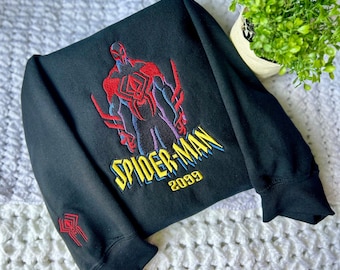 May include: Black sweatshirt featuring an embroidered Spider-Man 2099 design in red, blue, yellow, and purple. The design includes the Spider-Man figure and the text "SPIDER-MAN 2099". A smaller Spider-Man logo is embroidered on the sleeve.