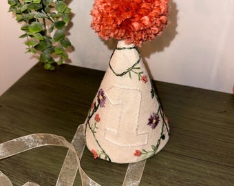 May include: A cream-colored party hat with the number "1" embroidered on the front. The hat is decorated with small, colorful flowers and green vines. A large, fluffy orange pom-pom sits atop the hat. A sheer, cream-colored ribbon is attached to the hat.