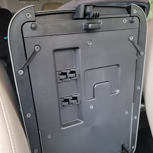 May include: A black plastic storage compartment with a hinged lid and multiple screws. The compartment has a rectangular opening and two small plastic clips on the inside.