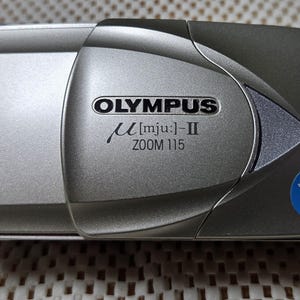 May include: Silver Olympus µ[mju:]-II Zoom 115 camera. The compact camera has a rectangular shape with rounded edges. The Olympus logo and model name are visible on the front. A blue "All Weather" sticker is on the bottom right corner.