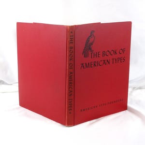 May include: A red hardcover book titled The Book of American Types by American Type Founders. The front cover features a black illustration of a bird and black text. The spine also displays the title in black text against the red background.