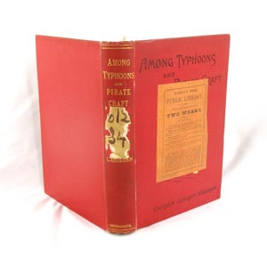 May include: A vintage book titled "Among Typhoons and Pirate Craft" with a red cover and spine. The spine features the title and author's name, with handwritten numbers. A library label is affixed to the cover.