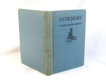 Puede incluir: Un libro abierto titulado "Astronomy" de Russell-Dugan-Stewart. El libro tiene una cubierta azul claro con el título en azul oscuro y una pequeña ilustración. El lomo es de color verde claro con texto.