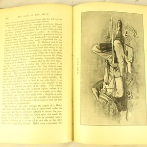 May include: An open book with text and a black and white illustration of a person holding a rifle. The illustration is on the right page, and the text is on the left page. The book has aged yellow pages.