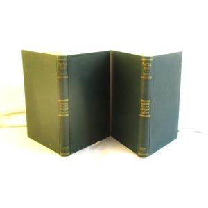 May include: Two dark green hardcover books are displayed, angled to create a V-shape. The spines feature gold lettering and decorative elements. The books appear to be vintage or antique, suggesting a literary or historical theme.
