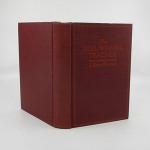 May include: A burgundy hardcover book with the title "The Soul-Winning Teacher" and the author "J.Riva Plummer" printed in gold lettering.