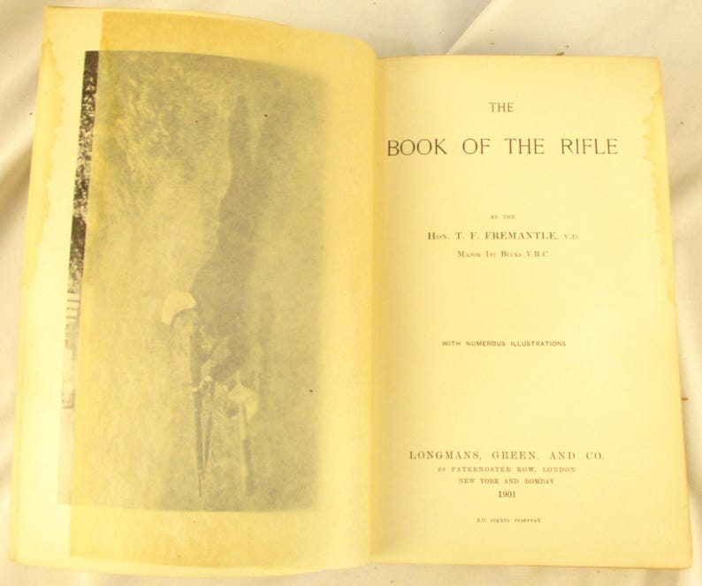 May include: An open book titled "THE BOOK OF THE RIFLE" with aged, yellowed pages. The left page features a black and white illustration. Published in 1901 by Longmans, Green, and Co. The text includes the author's name, H. T. F. Fremantle.