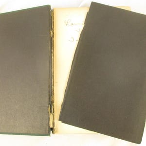 May include: An open antique book with a dark green cover on the left and a black cover on the right. The pages are off-white with handwritten script. The book's spine is visible, showing the binding.