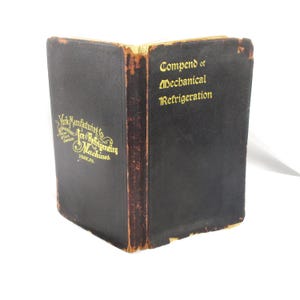 May include: A vintage book titled "Compend of Mechanical Refrigeration" with gold lettering on a black cover. The spine shows the title and the book's edge is worn. The cover also features the text "York Manufacturing Co. Ice & Refrigerating Machines, York, PA."