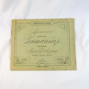 May include: A vintage, rectangular, light-tan instructional booklet titled Spencerian System of Penmanship, Common School Course. The cover features black decorative borders, ornate calligraphy, and the American Book Company imprint.