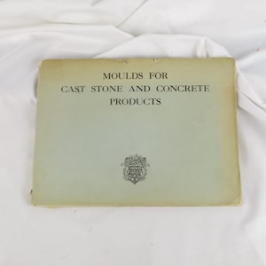 May include: A vintage book titled "Moulds for Cast Stone and Concrete Products" with text in black. The book has a light green cover with a decorative emblem at the bottom. The book is lying on a white surface.