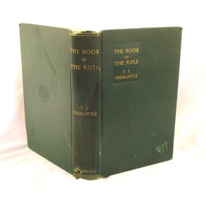 May include: An antique book, "THE BOOK OF THE RIFLE" by T.F. Fremantle, with a dark green cover and spine. Gold lettering is visible on the spine and cover. The book is housed in a dark green protective case.