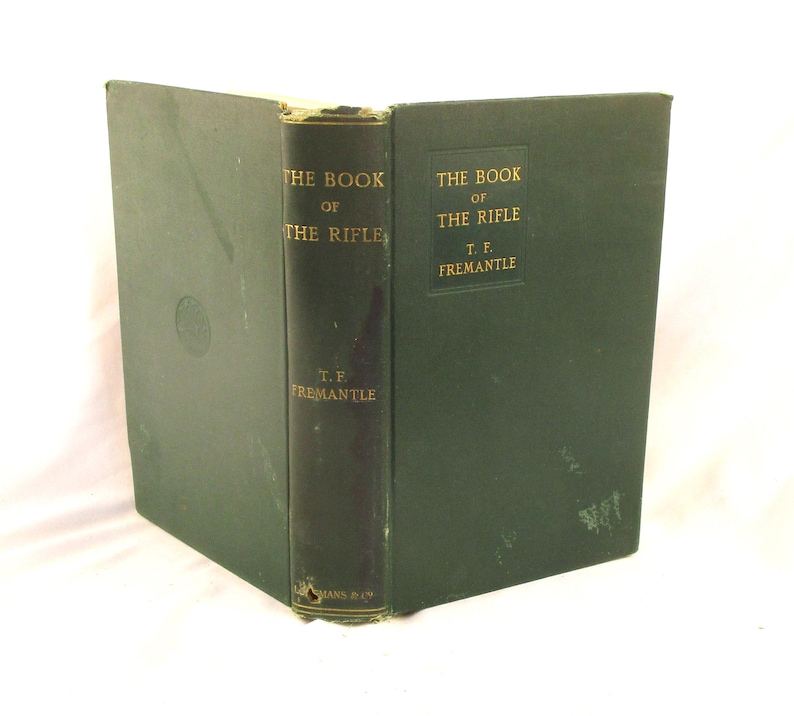 May include: An antique book, "THE BOOK OF THE RIFLE" by T.F. Fremantle, with a dark green cover and spine. Gold lettering is visible on the spine and cover. The book is housed in a dark green protective case.
