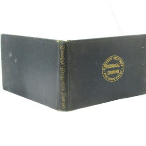 May include: An open, vintage book with a dark cover. The spine reads "Hawkins Mechanical Drawing." The front cover features a gold emblem with the text "Hawkins Self Help Mechanical Drawing For Home Study."