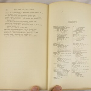 May include: An open book with aged, off-white pages. The left page lists titles under "THE BOOK OF THE RIFLE." The right page shows an index with various entries. The book is held open by two hands.