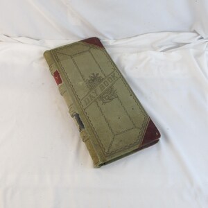 May include: An antique day book with a faded green cover and burgundy corners. The cover features the words "DAY BOOK" in a decorative font, along with ornate embellishments. The spine shows signs of wear, adding to its vintage charm.