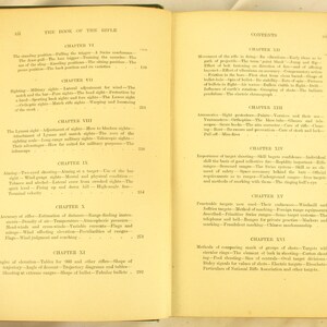 May include: An open book with aged, yellowed pages, titled "THE BOOK OF THE RIFLE." The left page lists chapters on rifle techniques, while the right page details contents, including target shooting and rifle mechanics. The book's cover is green.