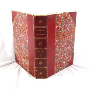 May include: Antique book titled "Constantinople" Volume 1, featuring a red leather spine with gold lettering and decorative emblems. The book's covers display a marbled pattern in red, blue, and gold, creating a vintage aesthetic.