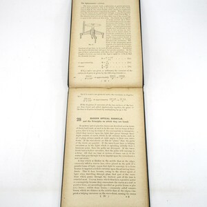 Peut inclure: Un livre vintage avec une couverture en cuir noir et des pages jaunies. Le livre est ouvert &agrave; une page avec un sch&eacute;ma d'une lentille et du texte sur les formules optiques modernes et les principes sur lesquels elles sont bas&eacute;es.
