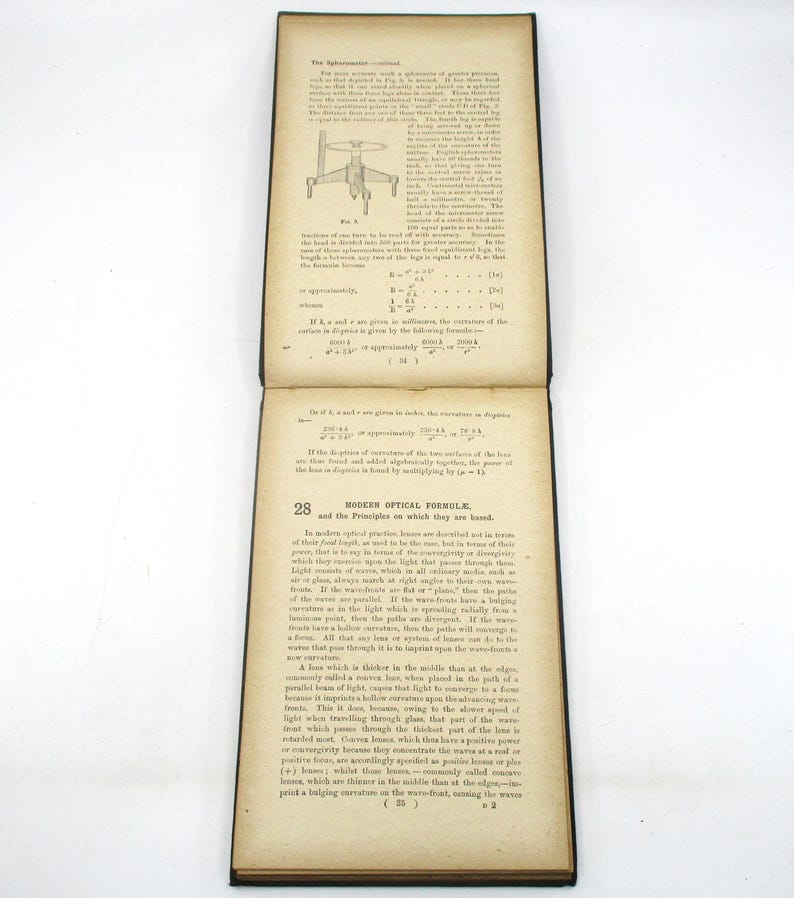 Peut inclure: Un livre vintage avec une couverture en cuir noir et des pages jaunies. Le livre est ouvert &agrave; une page avec un sch&eacute;ma d'une lentille et du texte sur les formules optiques modernes et les principes sur lesquels elles sont bas&eacute;es.