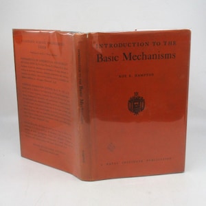 May include: An open book with a red cover titled "Introduction to the Basic Mechanisms" by Roy E. Hampton. The book has a glossy finish and features text on the spine and inside cover. A Naval Institute Publication is printed at the bottom.