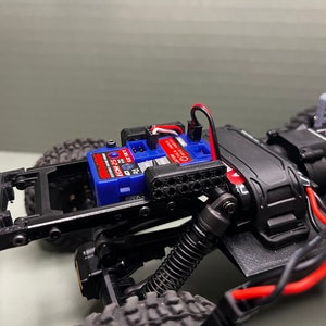 May include: A close-up of a black and gray RC car chassis with a blue and red electronic speed control (ESC) mounted on top. The ESC is labeled "Traxxas" and "ECM 25".