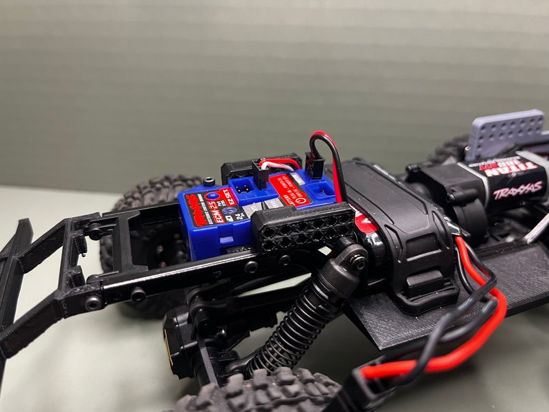 May include: A close-up of a black and gray RC car chassis with a blue and red electronic speed control (ESC) mounted on top. The ESC is labeled "Traxxas" and "ECM 25".