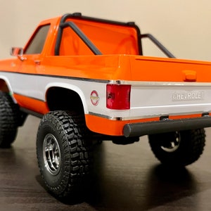 May include: Orange and white Chevrolet pickup truck with a black roll bar and black wheels. The truck has a white stripe along the side and the word "Chevrolet" on the tailgate.