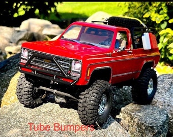 May include: A red Chevrolet K5 Blazer pickup truck with a black roll cage, a spare tire on the roof, and a black tube bumper. The truck is sitting on rocks and has large off-road tires.