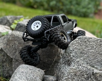 May include: A black and gray toy truck with large tires is driving over rocks. The truck has a roll cage and a black bed. The truck is a scale model of a real truck.