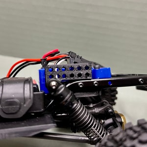 May include: Close-up of a black and blue RC car chassis with a black and red wire harness. The chassis has a black plastic body and a blue plastic battery holder. The battery holder has a black plastic grid on the front.
