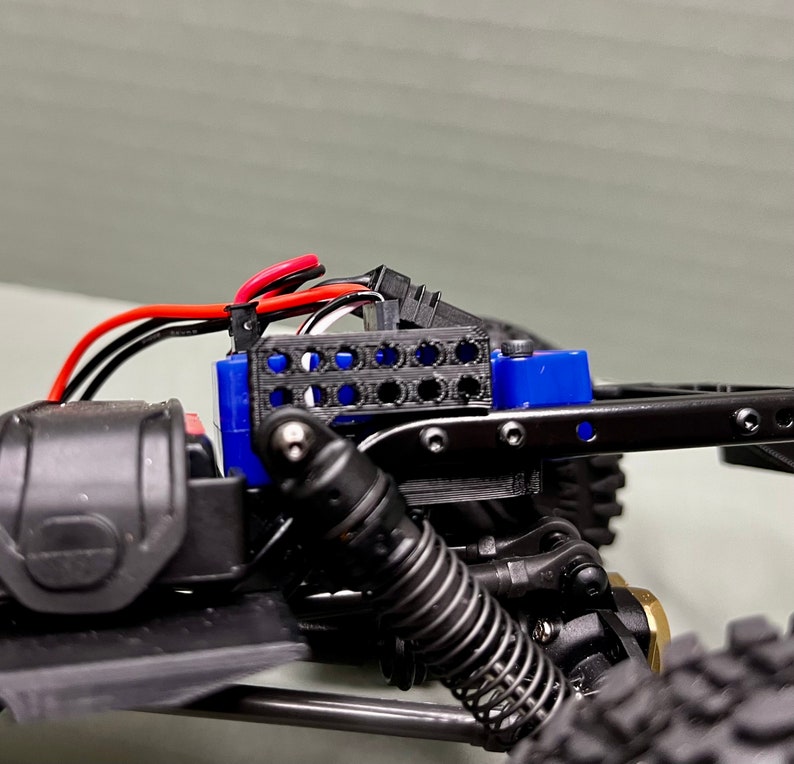 May include: Close-up of a black and blue RC car chassis with a black and red wire harness. The chassis has a black plastic body and a blue plastic battery holder. The battery holder has a black plastic grid on the front.