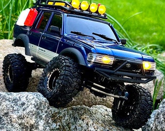 May include: A blue and silver toy truck with a black bumper and black tires. The truck has a white and red jerrycan on the roof and yellow lights on the front.