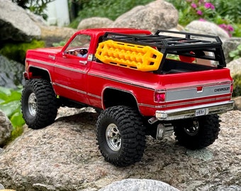 May include: A red Chevrolet pickup truck model with large off-road tires. It has a black rack on the bed and a yellow traction board. The truck is parked on a rocky surface.