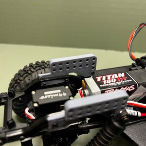 May include: Close-up of a black and gray RC car chassis with a gray plastic bumper and a Traxxas Titan 180 877 motor. The car has a black and gray servo and a red and white wire.