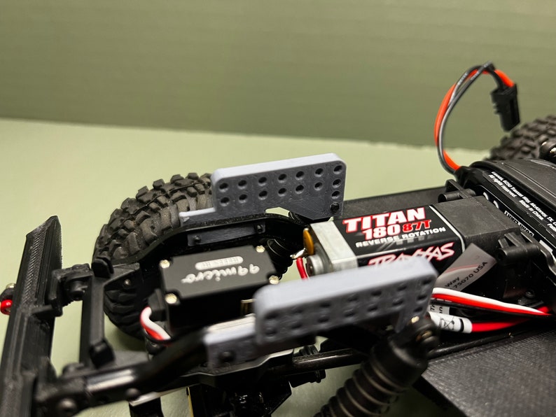May include: Close-up of a black and gray RC car chassis with a gray plastic bumper and a Traxxas Titan 180 877 motor. The car has a black and gray servo and a red and white wire.