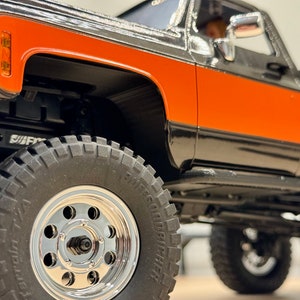 May include: A black and orange pickup truck with chrome wheels. The truck has a black bumper and a black step bar. The truck is sitting on a white surface.