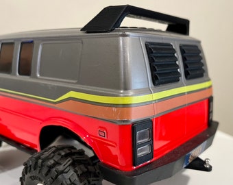 May include: A red, gray, and yellow toy van with a black roof rack and a black rear window. The van has a black bumper and a black tow hitch.