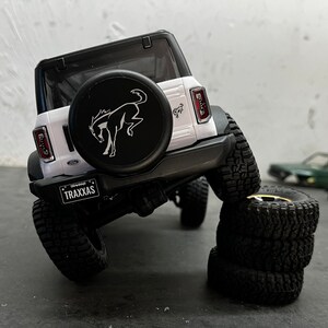 May include: White Ford Bronco model car with a black spare tire and a running horse logo. The car is on a gray surface with a stack of four black tires.