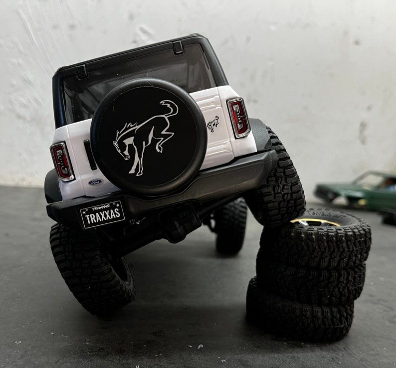 May include: White Ford Bronco model car with a black spare tire and a running horse logo. The car is on a gray surface with a stack of four black tires.
