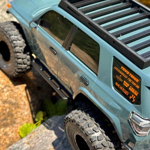 May include: A teal-colored toy SUV with black tires and a black roof rack. The vehicle has orange and white decals on the rear window. The SUV is positioned on a rocky surface, suggesting an off-road theme.