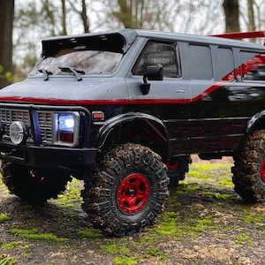 May include: Black and gray van with red accents, featuring a roof rack and off-road tires. The van has a black bumper with two round headlights and a grill.