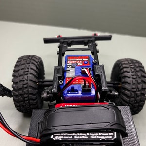 May include: A close-up of a black and red remote control car chassis with a blue electronic speed control (ESC) and a black battery pack. The car has black tires and a black frame.