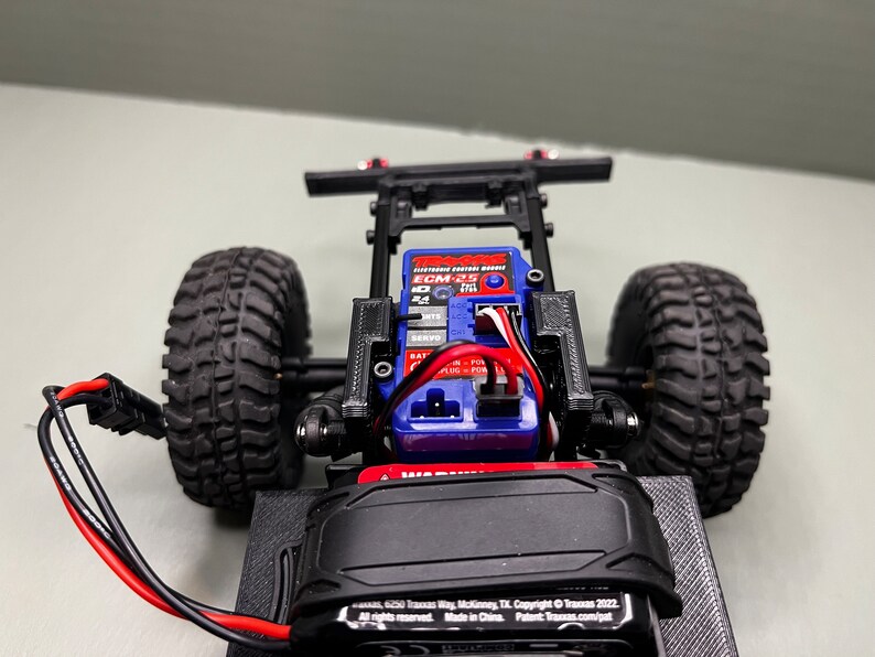 May include: A close-up of a black and red remote control car chassis with a blue electronic speed control (ESC) and a black battery pack. The car has black tires and a black frame.