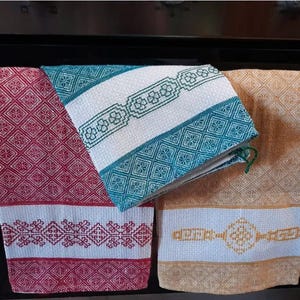 Puede incluir: Tres toallas de cocina con diferentes patrones. La primera toalla es roja con patrones de diamantes blancos y un borde blanco con un diseño bordado rojo. La segunda toalla es blanca con un patrón de diamantes azul y verde y un borde blanco con un diseño bordado verde. La tercera toalla es amarilla con un patrón de diamantes blancos y un borde blanco con un diseño bordado amarillo.