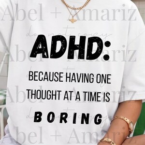 May include: A white short-sleeved t-shirt featuring black text that reads: ADHD: BECAUSE HAVING ONE THOUGHT AT A TIME IS BORING. The shirt has a crew neckline and a relaxed fit, with the bold, sans-serif lettering centered on the front chest area.