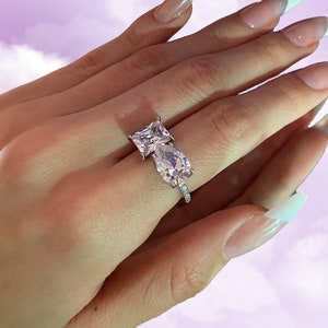 Könnte beinhalten: Ein silberner Ring mit zwei klaren Edelsteinen, einem quadratischen und einem tropfenförmigen Stein. Das Band ist mit kleinen Steinen besetzt. Der Hintergrund ist ein weiches Lila mit einem bewölkten Himmel.